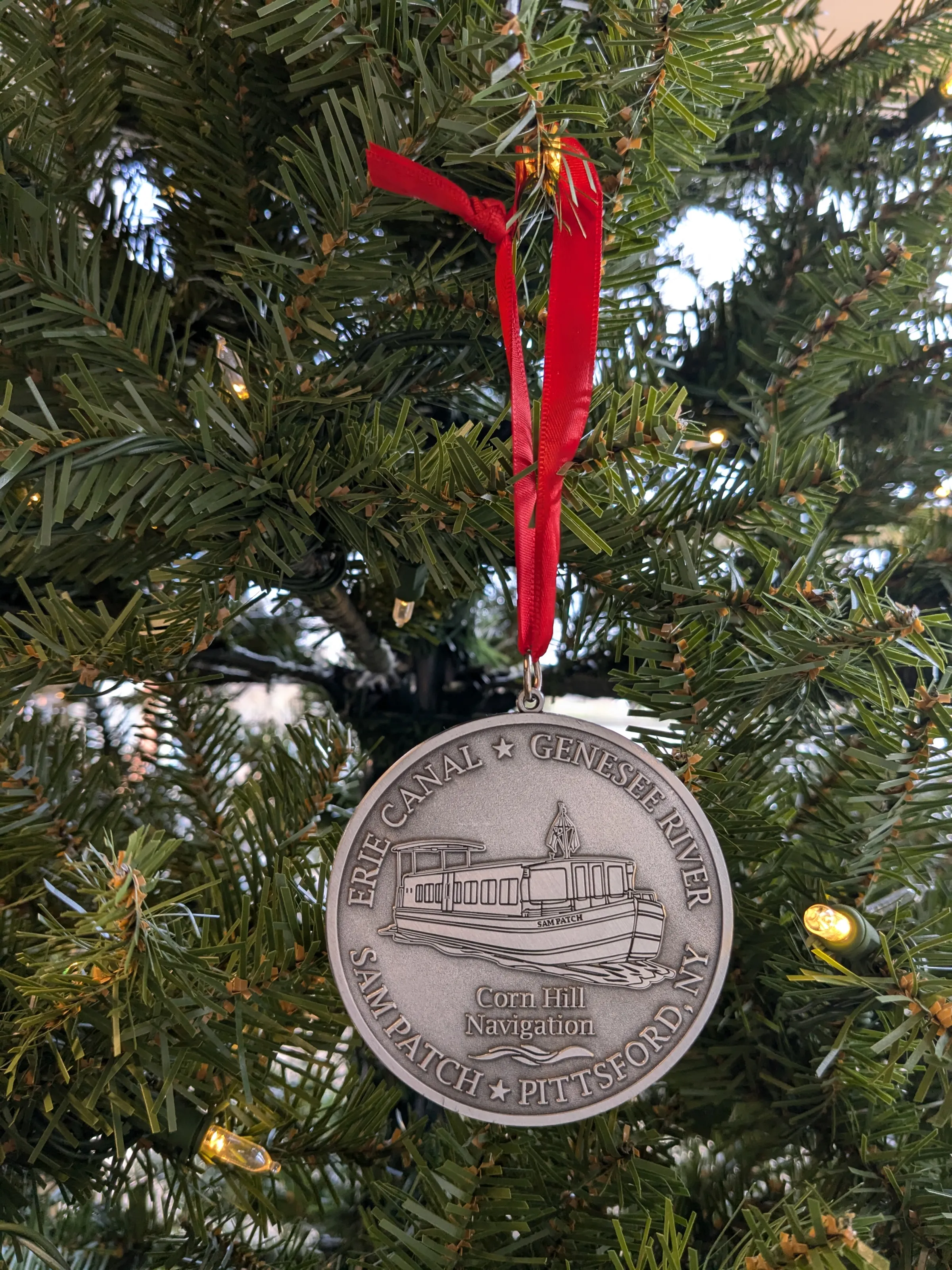 Metal ornament with a boat design on a Christmas tree, hung by a red ribbon.