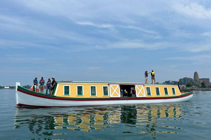 People standing on a colorful canal boat in calm water, with buildings in the background.