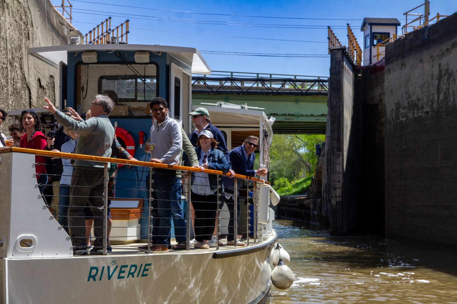 People on a boat named RIVERIE in a lock, with a bridge and greenery in the background.