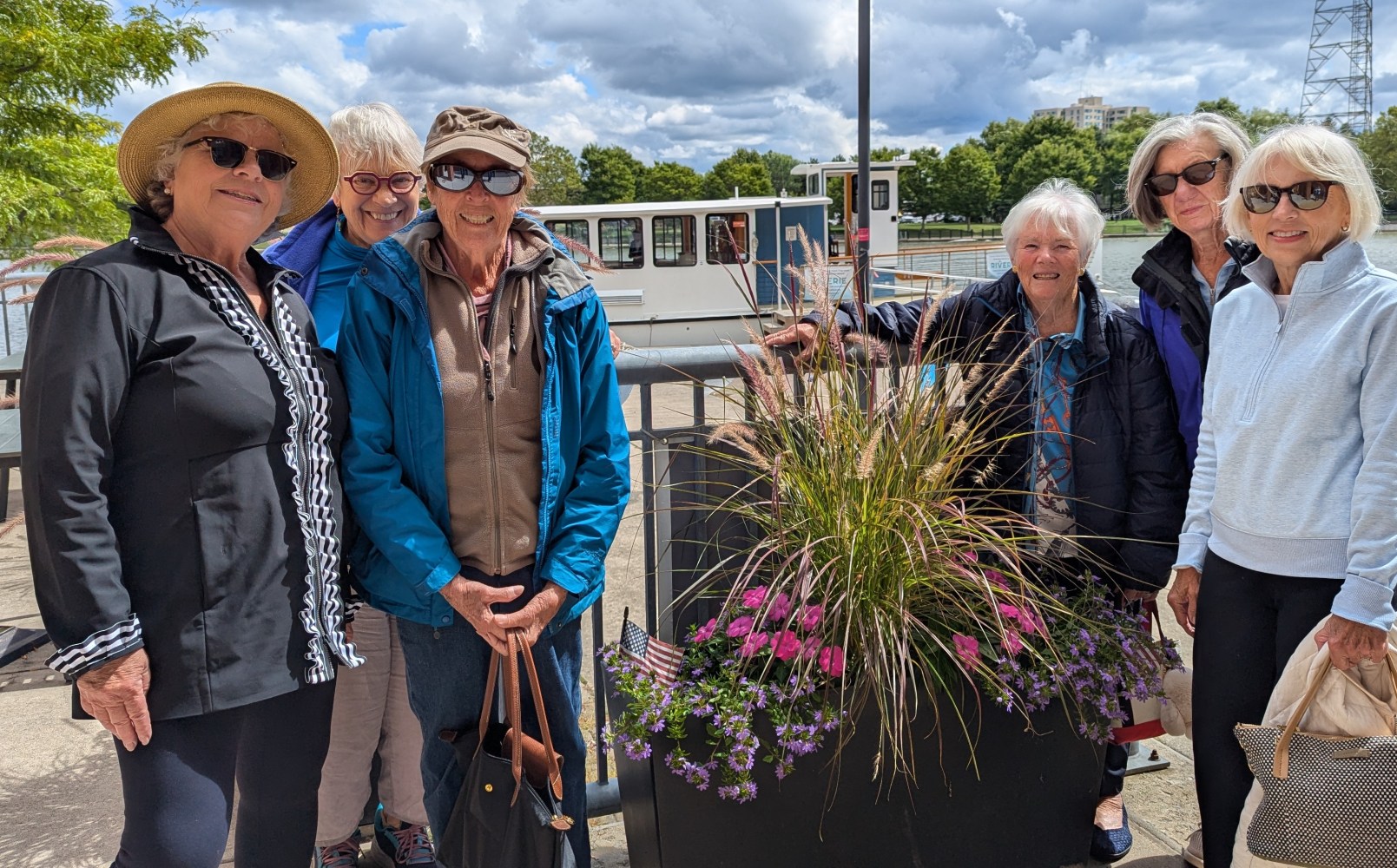 Six women smiling outdoors near a railing with flowers and a riverboat in the background.