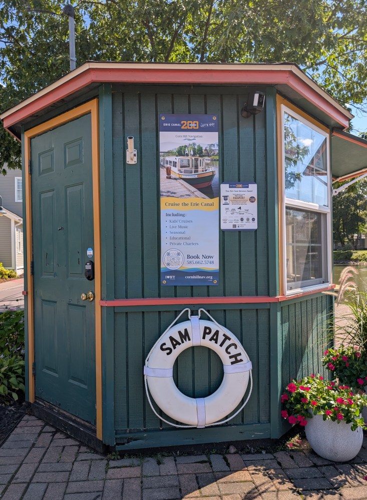 Small green kiosk with Erie Canal signage and Sam Patch lifesaver, surrounded by flowers.