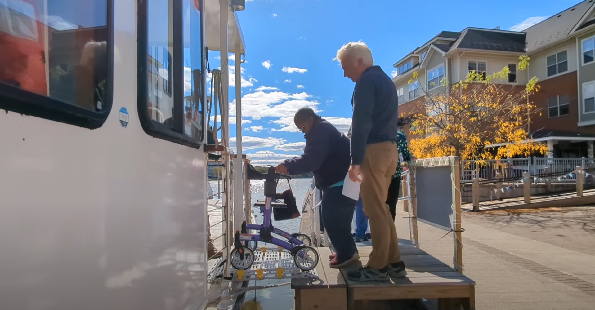 Two people boarding a boat using a ramp, one with a walker near a waterfront area on a sunny day.