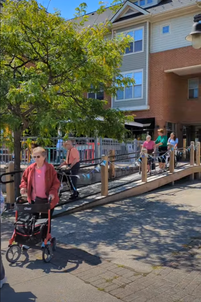 People using walkers on a wooden ramp outside a building with trees and sidewalk.
