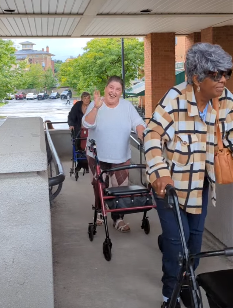 Three women using walkers, one waving, walk under a canopy on a rainy day.