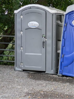 Gray portable toilet next to a blue one, outdoors on a gravel surface.